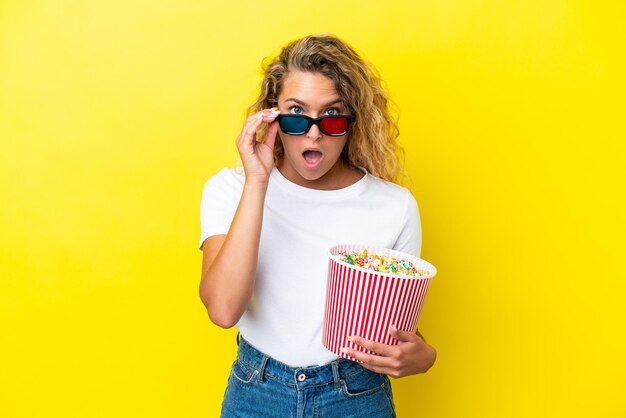 Mädchen mit lockigem Haar isoliert auf gelbem Hintergrund überrascht mit 3D-Brille und hält einen großen Eimer Popcorn