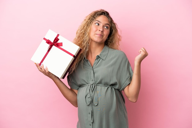 Mädchen mit lockigem Haar isoliert auf rosa Hintergrund schwanger und hält ein Geschenk, während sie Zweifel hat