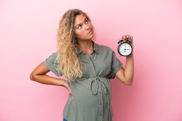 Mädchen mit lockigem Haar isoliert auf rosa Hintergrund schwanger und hält Uhr beim Denken