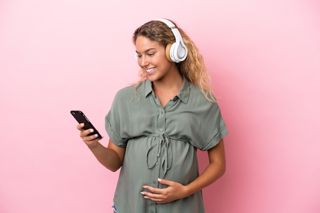 Mädchen mit lockigem Haar isoliert auf rosa Hintergrund schwanger und hört Musik mit einem Handy