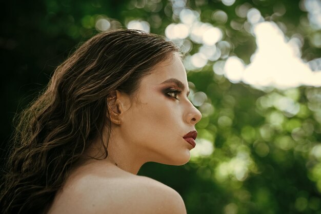 Mädchen mit Make-up-Gesicht, Schönheit. Frau mit langen brünetten Haaren auf Natur. Frau mit lockiger Frisur und nackter Schulter am Sommertag. Model mit Glamour-Look. Haarpflege, Therapie und Behandlung