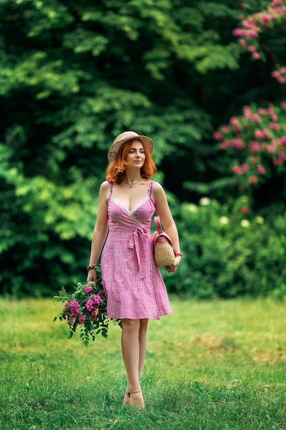 Mädchen mit roten Haaren in einem Hut in einem rosa Kleid mit einer Tüte Blumen draußen