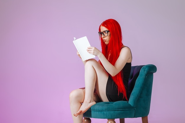 Mädchen mit roten langen Haaren sitzt auf einem Stuhl und liest ein Buch auf rosafarbenem Hintergrund