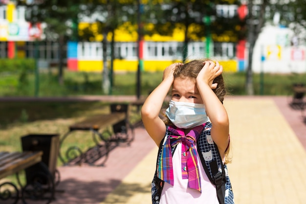 Mädchen mit Rucksack in der Nähe der Schule nach dem Unterricht nimmt medizinische Maske ab, unglücklich, müde und Kopfschmerzen