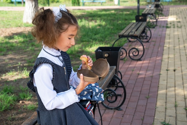 Mädchen mit Rucksack isst in einer Sandwichbox verpacktes Sandwich in der Nähe der Schule, ein schneller Snack mit einem Brötchen, ungesundes Essen, Mittagessen aus der Schule, zurück zur Schule, Bildung, Grundschulklassen, 1. September