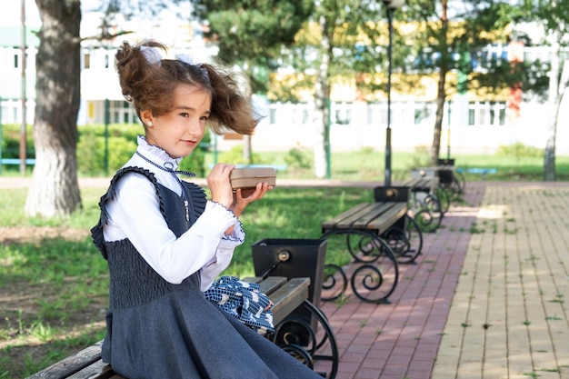 Mädchen mit Rucksack isst in einer Sandwichbox verpacktes Sandwich in der Nähe der Schule, ein schneller Snack mit einem Brötchen, ungesundes Essen, Mittagessen aus der Schule, zurück zur Schule, Bildung, Grundschulklassen, 1. September