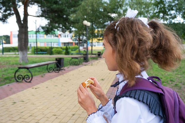 Mädchen mit Rucksack isst Kuchen in der Nähe der Schule, einen schnellen Snack mit einem Brötchen, ungesundes Essen, Mittagessen aus der Schule, zurück zur Schule, Bildung, Grundschulklassen, 1. September