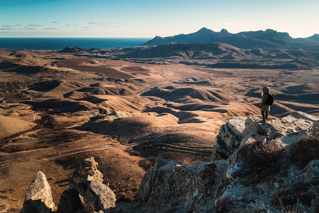 Mädchen mit Rucksack steht am Rand einer Klippe in den Bergen mit Blick auf das Tal bei Sonnenuntergang, Blick von hinten.