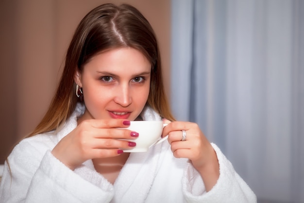 Mädchen mit slawischem Aussehen im Mantel mit weißer Tasse sitzt am Tisch. Weiblich nimmt die Emotionen des Schauspielers. Junge charmante Frau sitzt im Hotelzimmer und trinkt Kaffee oder Tee. Entspannungskonzept