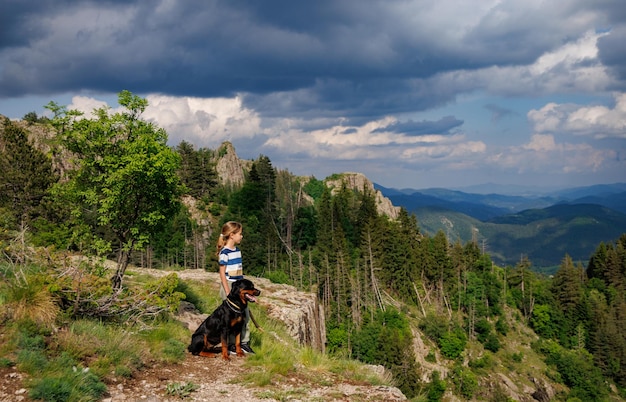 Mädchen mit steht neben ihrem Hund der Rottweiler-Rasse auf einem Gipfel mit Vegetation gegen den bewölkten Himmel