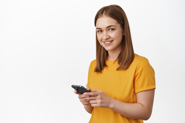 Mädchen mit Telefon. Junge blonde Frau mit Handy, SMS-Nachricht, lächelnd und glücklich in die Kamera schauend, im gelben T-Shirt vor weißem Hintergrund stehend.