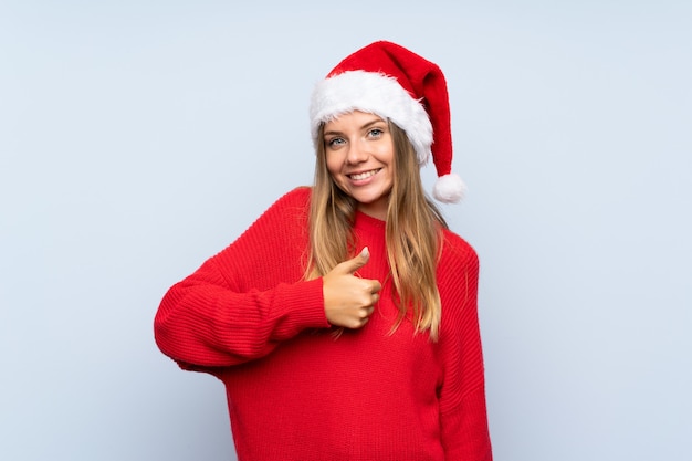 Mädchen mit Weihnachtshut über dem lokalisierten blauen Wandgeben Daumen up Geste