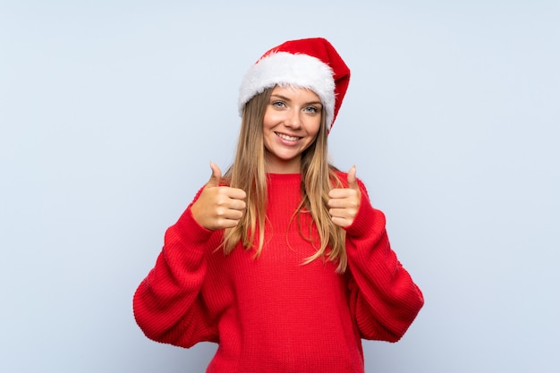 Mädchen mit Weihnachtshut über dem lokalisierten blauen Wandgeben Daumen up Geste