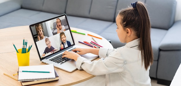 Mädchen schaut auf Laptop mit Videokonferenz-Kinderklassenkameraden, die auf dem Tisch stehen.