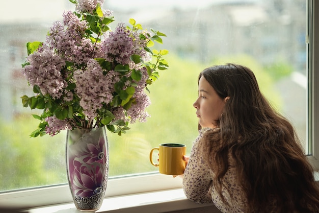 Mädchen schaut sich die Blumen an und trinkt Tee oder Kaffee aus einem gelben Becher daneben auf der Fensterbank steht eine Vase mit einem Fliederstrauß