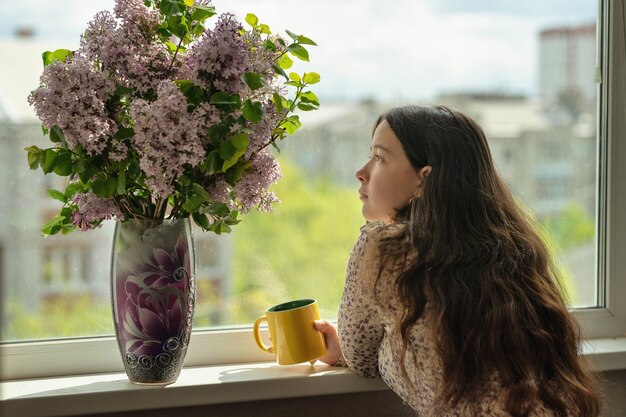 Mädchen schaut sich die Blumen an und trinkt Tee oder Kaffee aus einem gelben Becher daneben auf der Fensterbank steht eine Vase mit einem Fliederstrauß