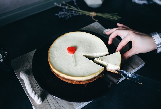 Mädchen schneidet mit klassischem Vanille New Yorker Käsekuchen des Messers auf dunkelvioletter Platte auf Holztablett mit Seilgriffen. Kleines rotes Herz liegt auf dem Dessert. Perfektes Hygge-Geschenk zum Valentinstag. Hausgemacht.