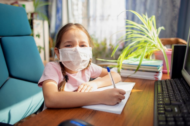 Mädchen Schulmädchen in einer medizinischen Maske macht Hausaufgaben vor einem Computer. Quarantäne und Heimunterricht