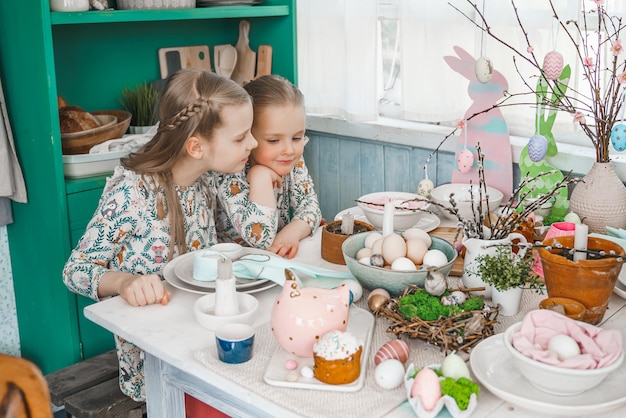 Mädchen Schwestern am Tisch mit Osterdekoration Feier in der Küche Tischlandschaft für zu Hause Osterferien Familie religiöse traditionelle festliche Mahlzeit Farbige Eierkuchen lustiger Hase Süßigkeiten süß