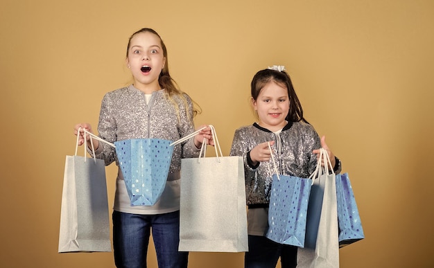 Foto mädchen schwestern freunde mit einkaufstüten beige hintergrund einkaufen und einkaufen schwarzer freitag verkauf und rabatt einkaufstag kinder halten bündel pakete kindermode erwarten sie mehr, zahlen sie weniger