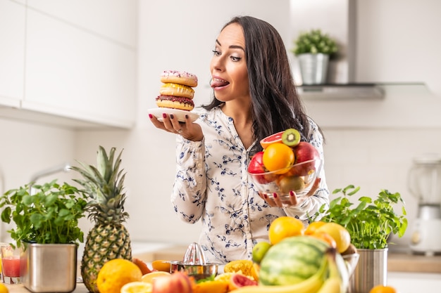 Mädchen sehnt sich nach ungesunden Donuts über gesundem Obstteller in einer Küche voller Früchte der gesunden Ernährung.