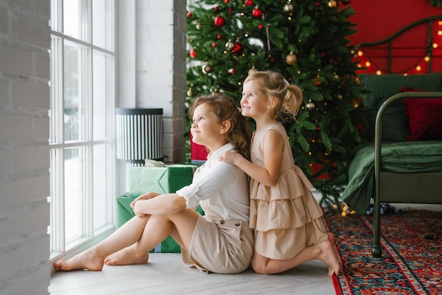 Mädchen sitzen am Fenster vor dem Hintergrund eines Weihnachtsbaums im Schlafzimmer, schauen aus dem Fenster und träumen von Magie