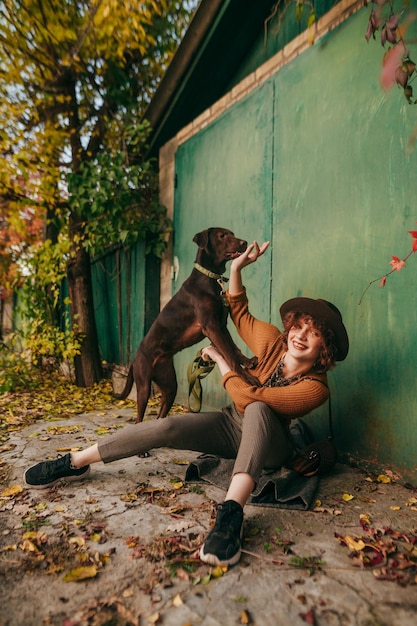 Mädchen sitzt am Herbsttag unter der grünen Wand des Landhauses und spielt mit einem Hund mit einem Lächeln im Gesicht