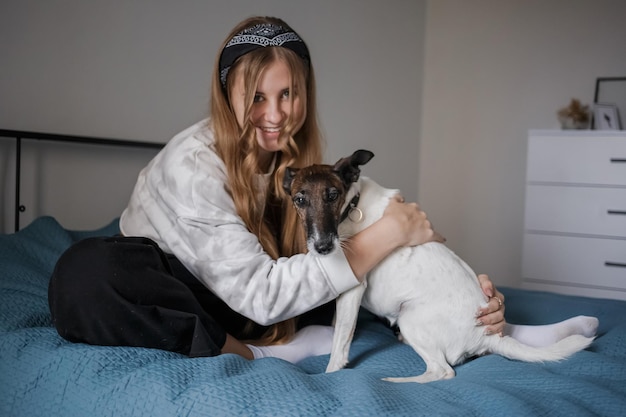 Mädchen sitzt auf einem Bett in ihrem Wohnzimmer und umarmt einen kleinen weißen Hund, der geradeaus schaut und lächelt