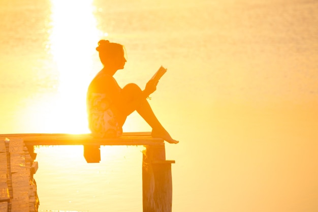 Foto mädchen sitzt auf einem pier und liest bei sonnenuntergang