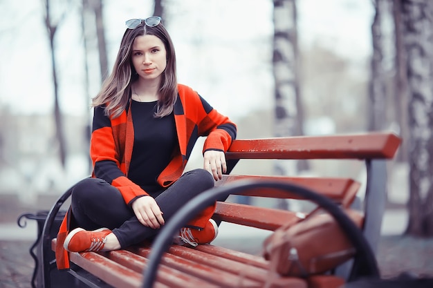 Mädchen sitzt auf einer Gartenbank / schönes Model posiert im Mantel auf Bank im Stadtpark sitzend, schönes Mädchen