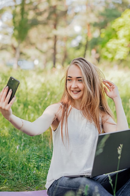 Mädchen sitzt im Freien und arbeitet an einem Laptop macht ein Selfie am Telefon freiberufliche Selbstbildung das Konzept des Fernlernens und der Arbeit im Freien