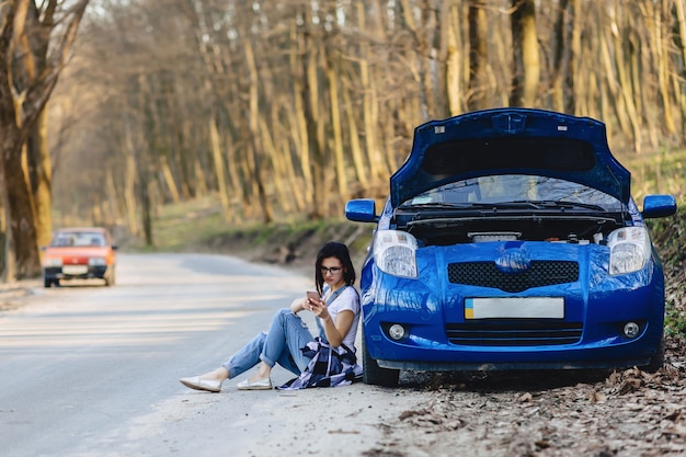 Mädchen sitzt neben kaputtem auto mit offener motorhaube und spricht telefonisch