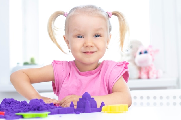 Mädchen spielt kinetischen Sand in Quarantäne. Blondes schönes Mädchen lächelt und spielt mit lila Sand auf einem weißen Tisch.