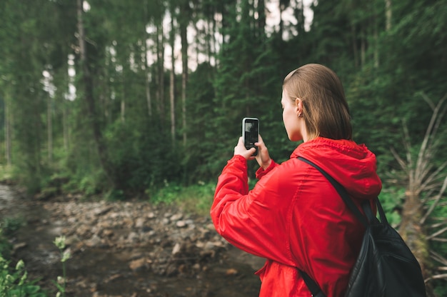 Mädchen steht im Wald nahe Gebirgsfluss und macht Foto der Landschaft auf Smartphone