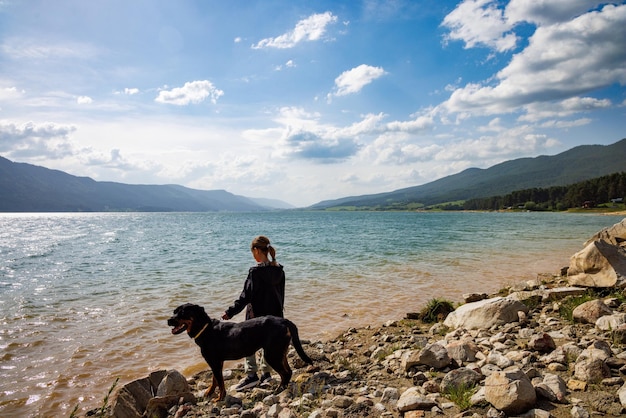 Mädchen steht in der Nähe des Hundes der Rottweiler-Rasse am Ufer in der Nähe des Sees vor dem Hintergrund der mit Wald bedeckten Bergkette