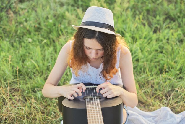 Foto mädchen teenager in hut spielt gitarre sitzen auf dem gras