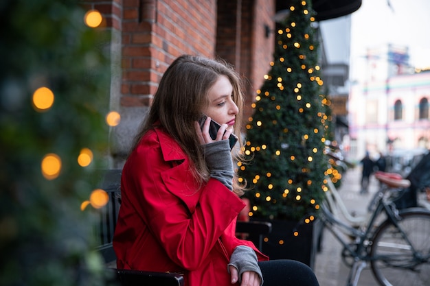 Mädchen telefoniert auf einer Bank in der Nähe von Weihnachtsbäumen, die mit Lichtern geschmückt sind