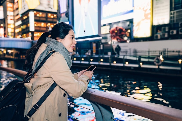 Mädchen Tourist stehen neben dem Fluss in Dotonbori Osaka Japan entspannen. Schöne Reisende, die ein Smartphone mit einer Online-SMS-Nachricht hält und die Nachtansicht des Kanals und der geschäftigen Stadt sieht.