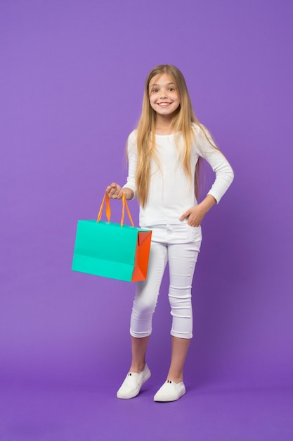 Mädchen tragen weißen Pullover und Hose isoliert auf lila Hintergrund Kind mit fröhlichem Gesicht Holding Papiertüte Kind überrascht von Geschenk-Geburtstag-Konzept Mädchen mit großem Lächeln beim Einkaufen