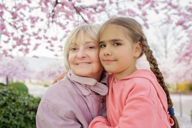 Mädchen umarmt ihre Großmutter in der Nähe eines blühenden Zweigs von Sakura beim Gehen im Frühlingsgarten