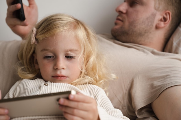 Foto mädchen und erwachsener mann liegen zu hause im bett und benutzen die telefone. kind und vater schauen sich geräte an, die zeit verbringen.