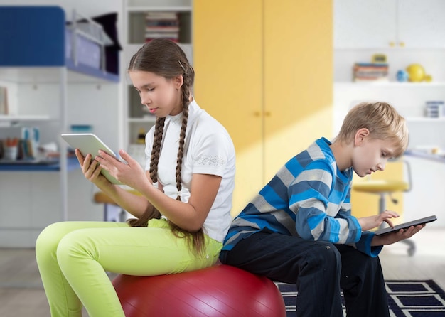 Mädchen und Junge sitzen auf einem großen Gummiball und schauen auf Pad Tablet PC-Bildschirme