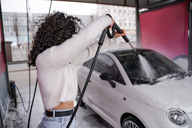 Mädchen waschen persönliches Auto auf SB-Autowäsche. Konzentrieren Sie sich auf den Vordergrund der lockigen Frau, die das Auto aus dem Schlauch wässert. Auto mit Seife und Wasser bedeckt