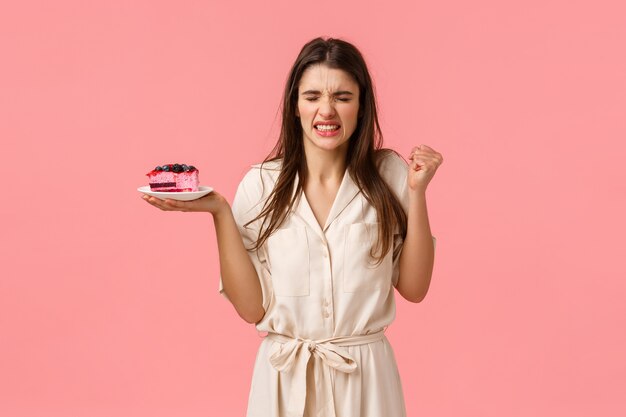 Mädchen wird von ekelhaftem Kuchengeschmack kotzen. Enttäuschter Versuch der jungen Frau, Nachtisch zu essen und Gesicht zu verziehen, vom schrecklichen Geruch zu schielen, Nachtisch zu halten und rosa Wand zu stehen