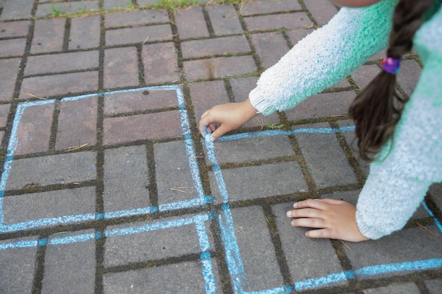 Mädchen zeichnet ein Himmel und Hölle mit Kreide auf dem Bürgersteig Kind beim Spielen des Spiels draußen Kind mit Pullover beim Spielen von Himmel und Hölle Aktivitäten für Kinder Kindermalereien auf Asphalt