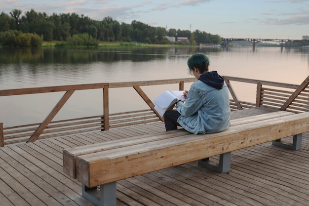 Mädchen zeichnet in einem Skizzenbuch beim Sitzen auf der Böschung im Sommer