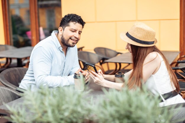 Mädchen zeigt ihrem Freund den Bildschirm eines Smartphones, während sie in einem Café im Freien sitzen.