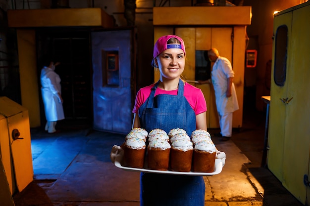 Mädchenbäcker hält ein Tablett mit heißem Gebäck in der Bäckerei. Sie trägt ein Jeanskleid und eine Mütze. Herstellung von Backwaren. Rack mit frischem knusprigen Gebäck.