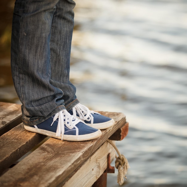 Mädchenbeine in blauen Turnschuhen, die auf einem Pier vor dem Hintergrund eines Sees oder eines kleinen Flusses stehen