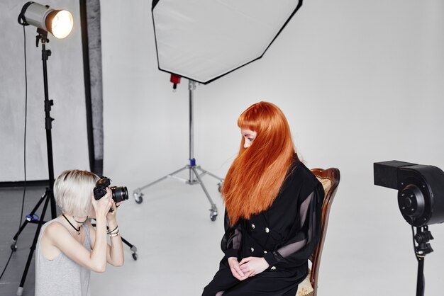 Foto mädchenfotograf, der modemodell in schwarz auf einem stuhl auf weißem hintergrund im studio fotografiert